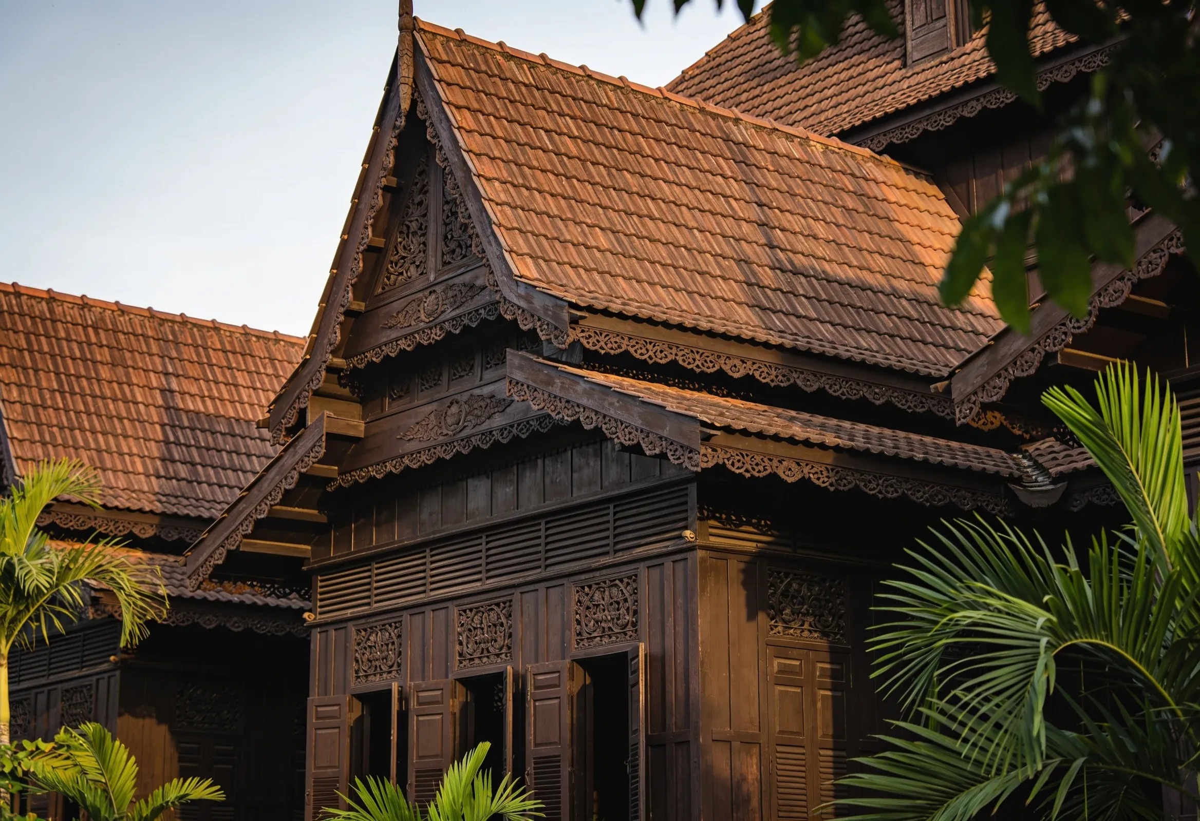 Morning light on the dark hand-carved timber facade and steep roof of Istana Jahar palace in Kota Bharu, a visitor in modest dress walking past at a distance, no identifiable faces in focus.